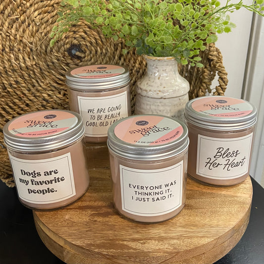 Five candles with humorous labels on a wooden surface, surrounded by decorative elements.