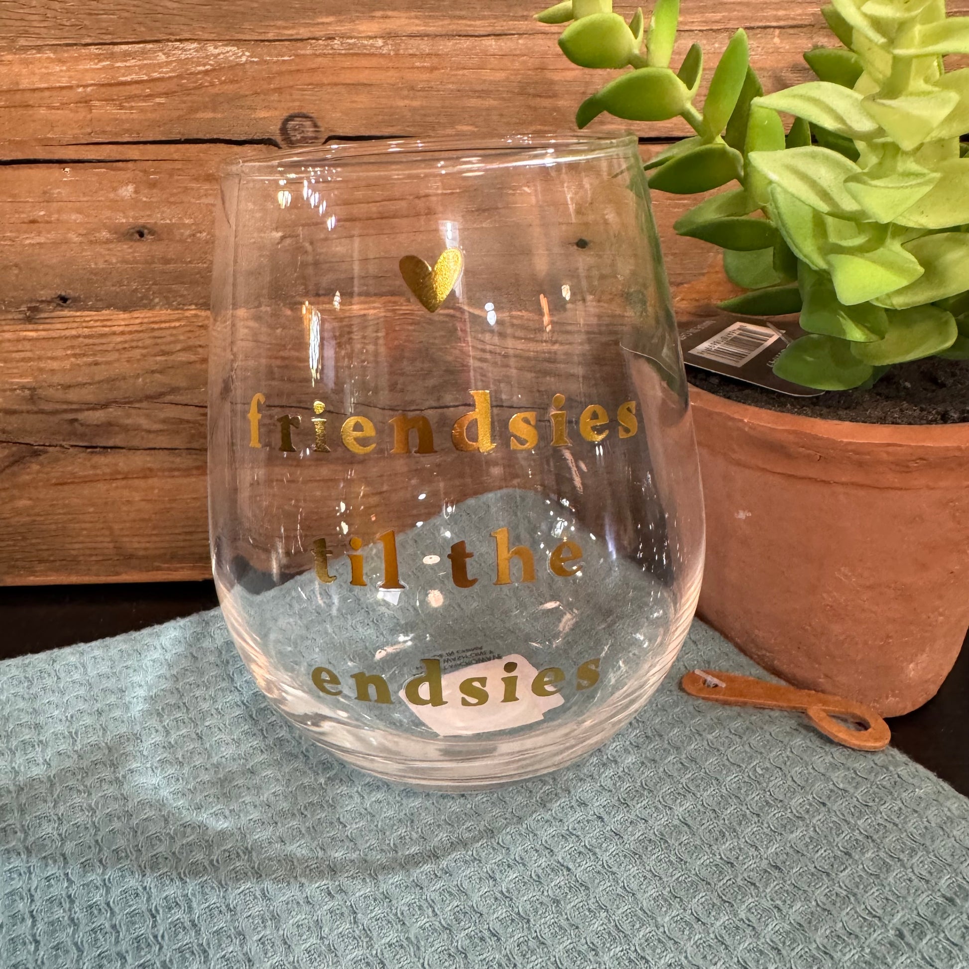 A clear stemless wine glass with gold lettering that reads 'friendsies til the endsies' and a small gold heart, placed on a textured light blue cloth with a wooden wall and potted succulent in the background.