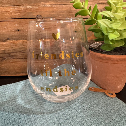 A clear stemless wine glass with gold lettering that reads 'friendsies til the endsies' and a small gold heart, placed on a textured light blue cloth with a wooden wall and potted succulent in the background.
