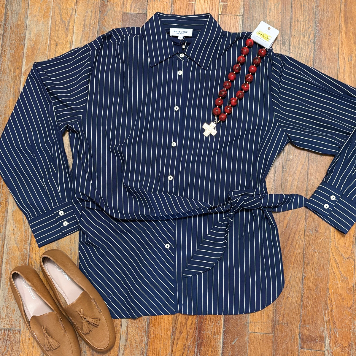 A navy blue long-sleeve button-up shirt with thin white vertical pinstripes, styled in a flat lay on a wooden floor with a brown tassel loafer and a red beaded necklace.