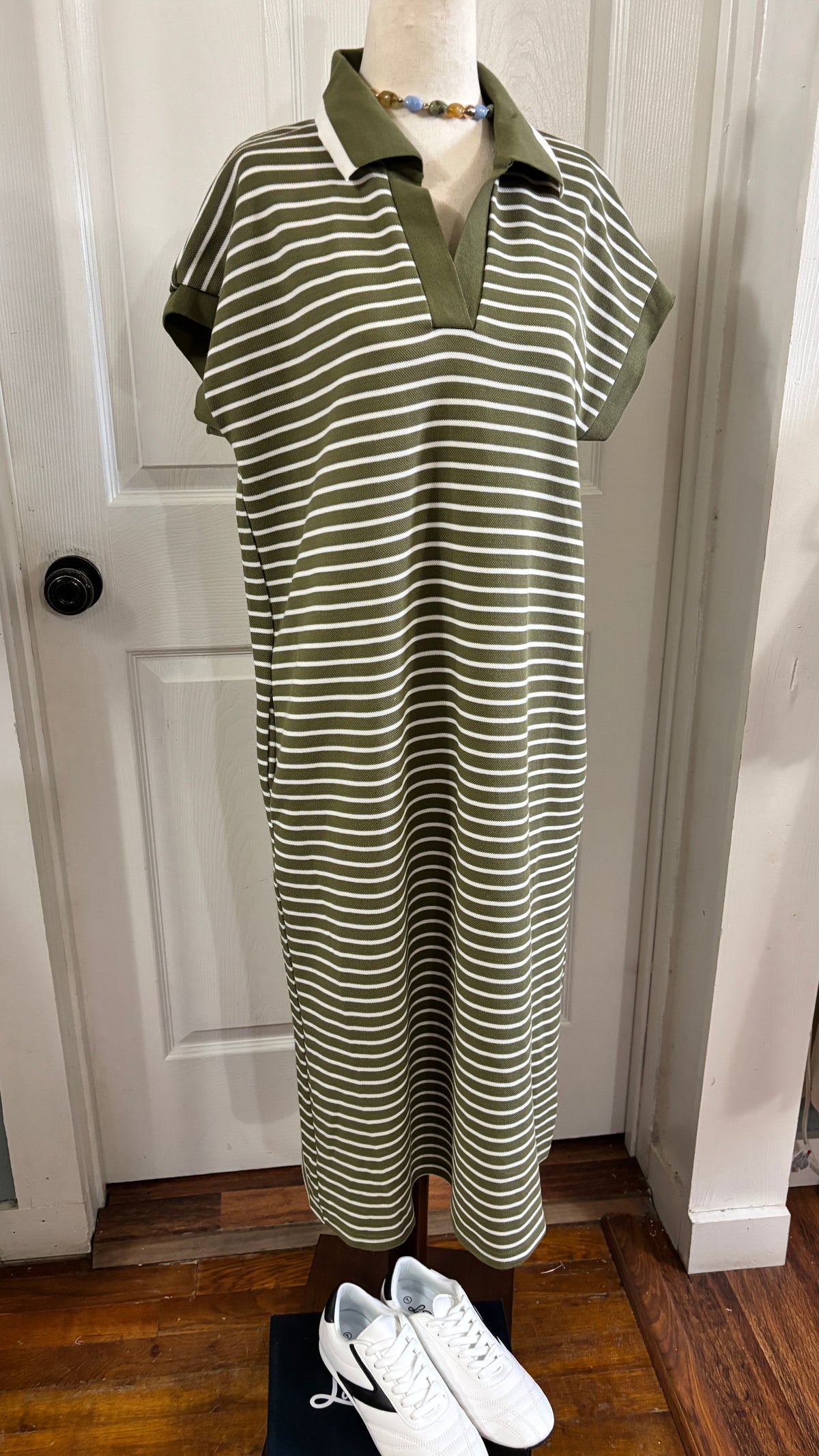 A short-sleeve polo dress with horizontal olive green and white stripes, displayed on a mannequin in front of a white door. White sneakers are placed at the bottom of the stand.