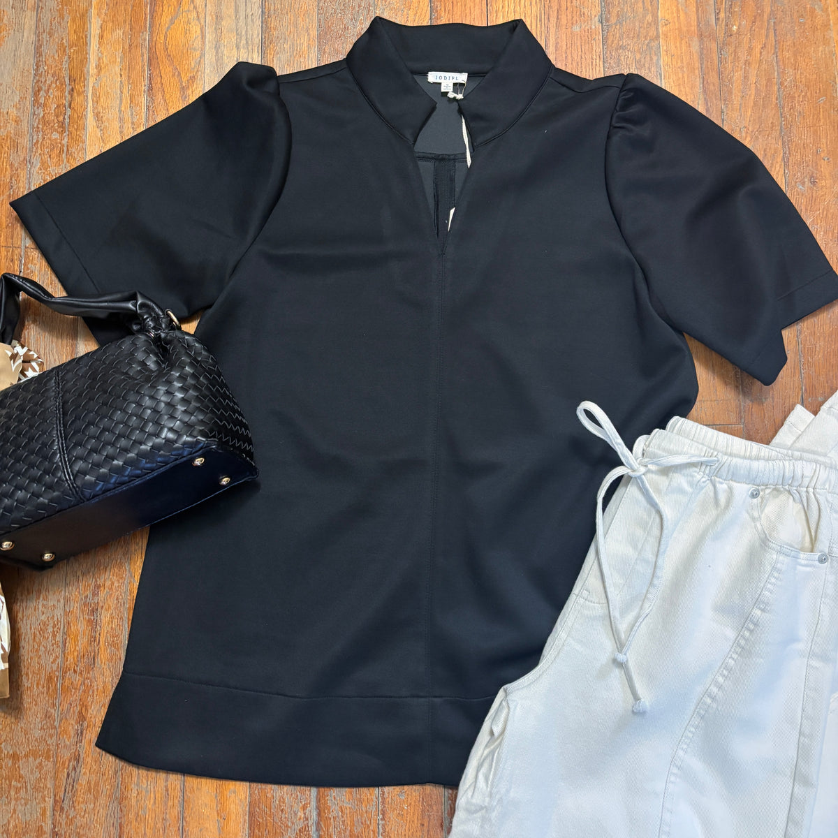 A black short-sleeve collared blouse with a half-zip front and structured shoulders, laid flat on a wooden floor alongside a black woven handbag and light gray drawstring pants.