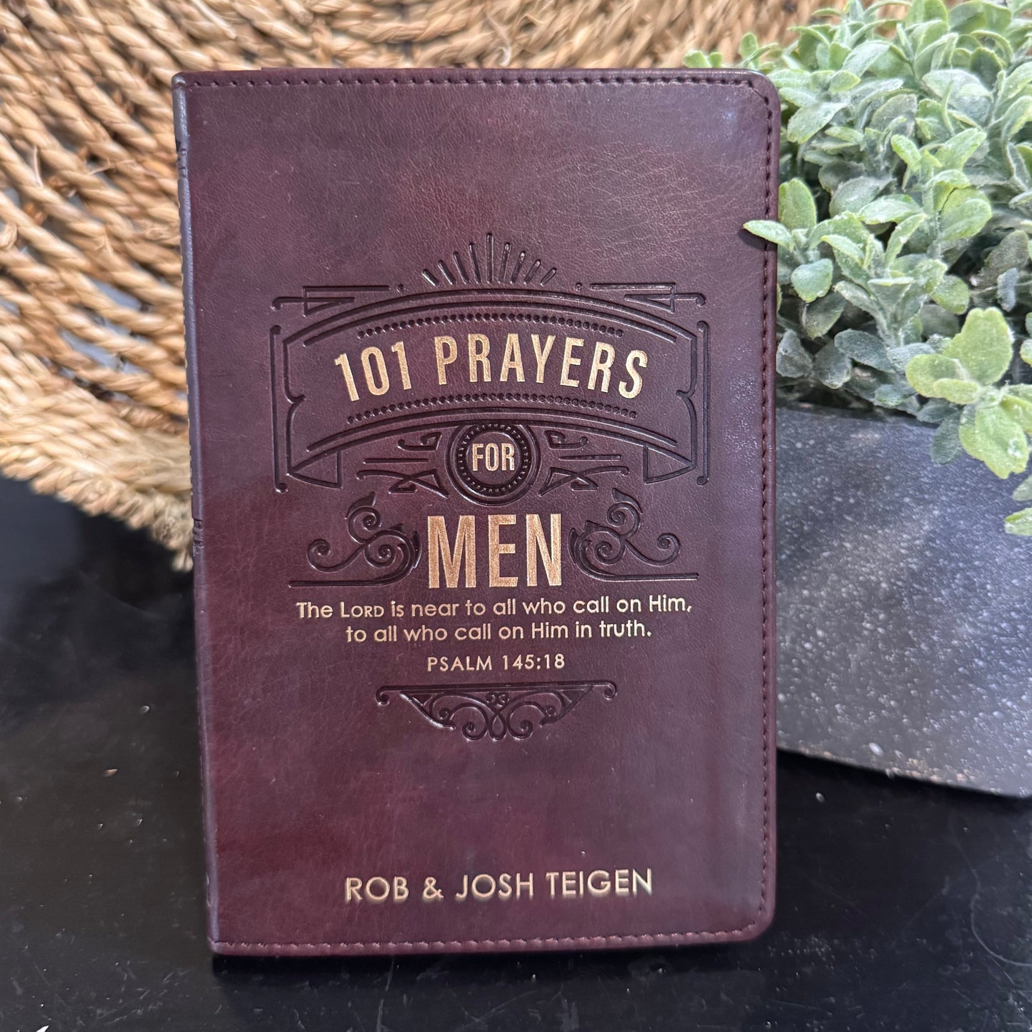 Brown leather book titled '101 Prayers for Men' by Rob & Josh Teigen on a dark surface with a plant in the background.