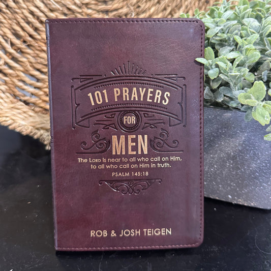 Brown leather book titled '101 Prayers for Men' by Rob & Josh Teigen on a dark surface with a plant in the background.