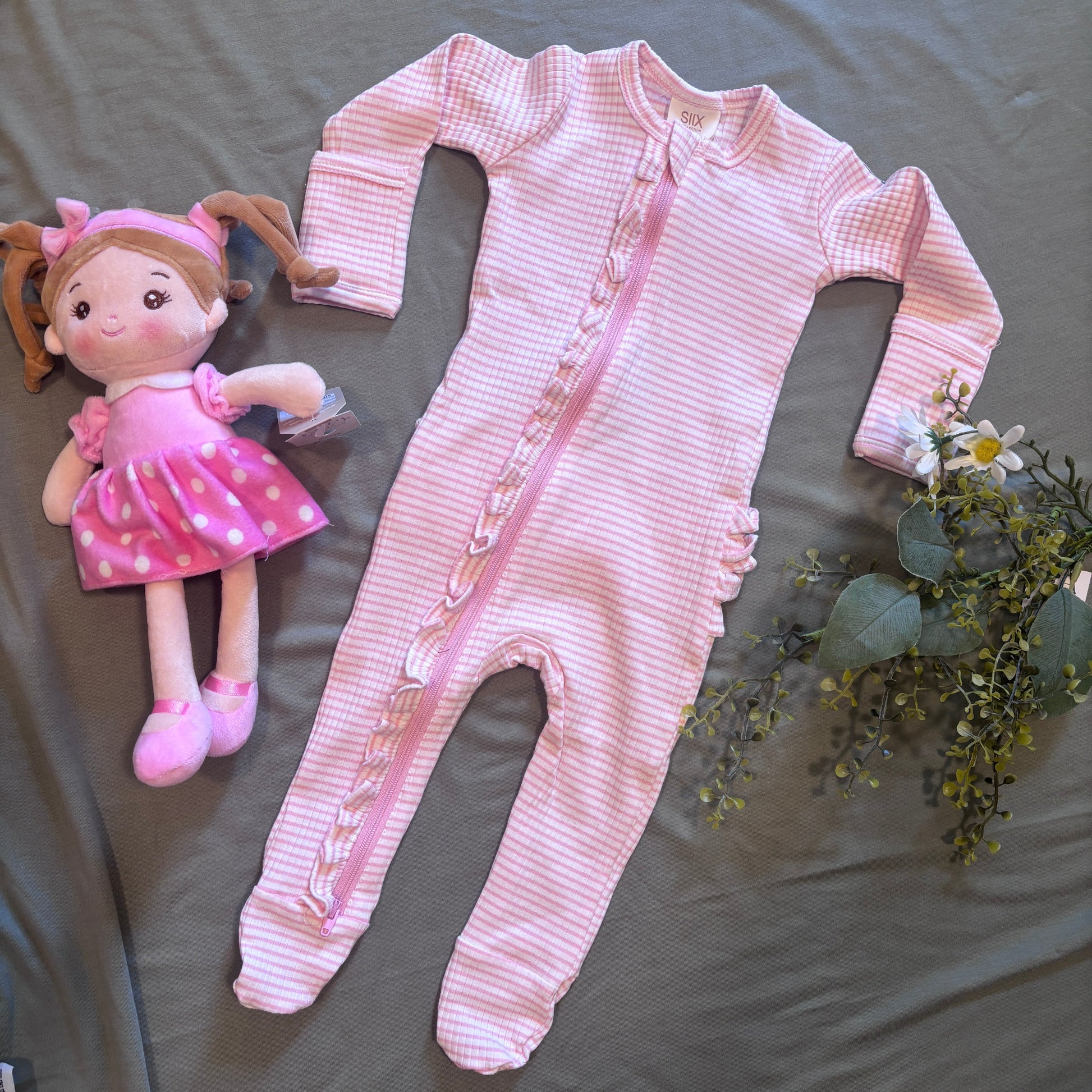 A pink and white gingham long-sleeve baby footed romper with a decorative braided zipper trim, laid flat on a gray fabric background next to a pink and white polka dot doll and green floral stems.