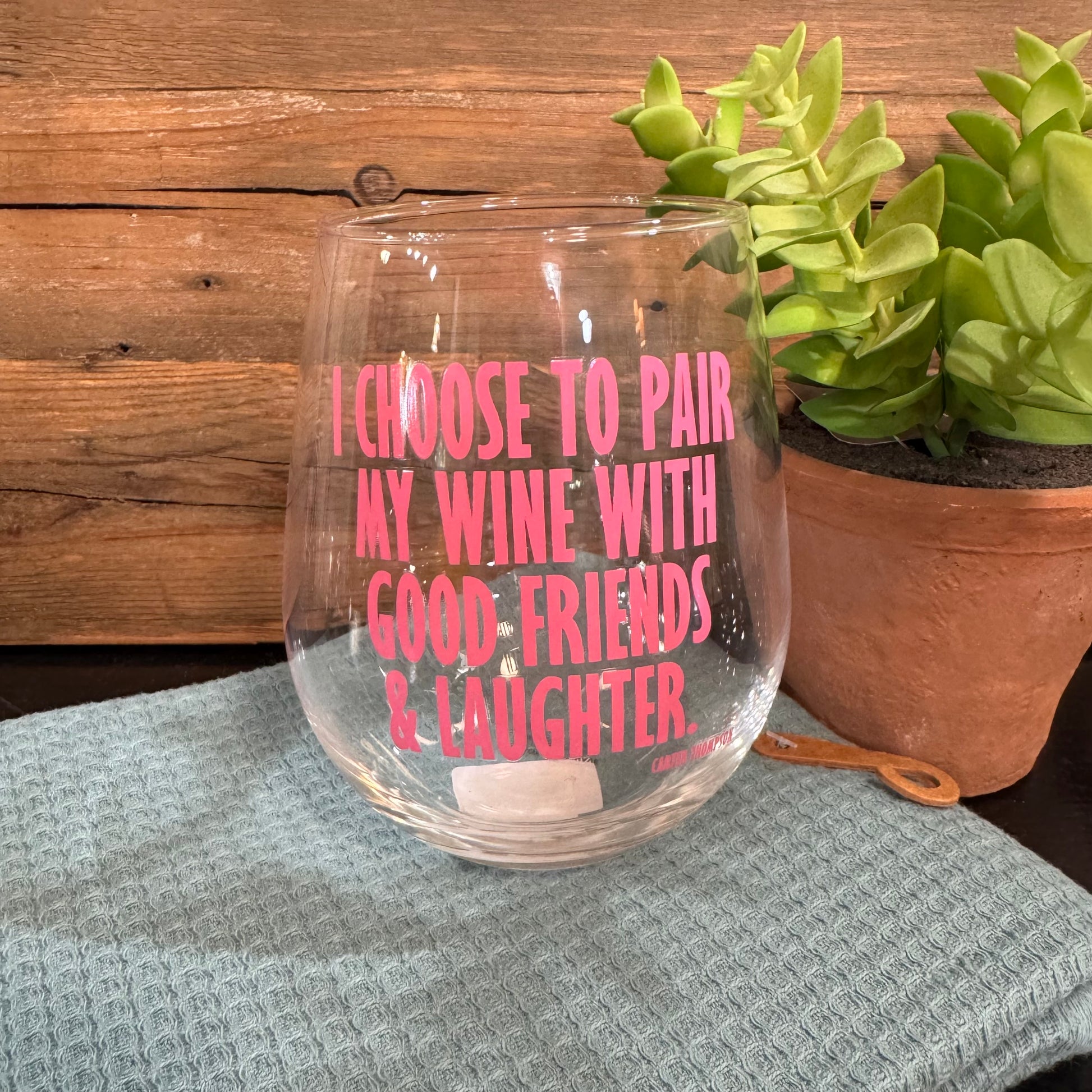 A clear stemless wine glass with pink text reading 'I CHOOSE TO PAIR MY WINE WITH GOOD FRIENDS & LAUGHTER' sits on a textured light blue cloth, with a wooden wall behind and a potted succulent to the right.
