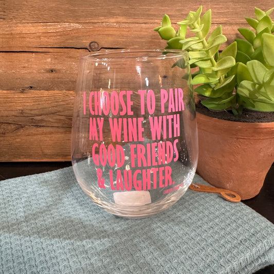 A clear stemless wine glass with pink text reading 'I CHOOSE TO PAIR MY WINE WITH GOOD FRIENDS & LAUGHTER' sits on a textured light blue cloth, with a wooden wall behind and a potted succulent to the right.
