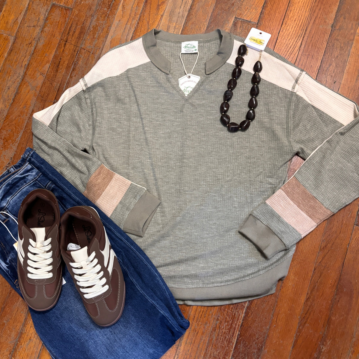 A flat-lay outfit arrangement on a wooden floor featuring a heather gray long-sleeve colorblock shirt with beige and brown panels, folded blue jeans, brown sneakers with white laces, a dark beaded necklace, and a small white tag.