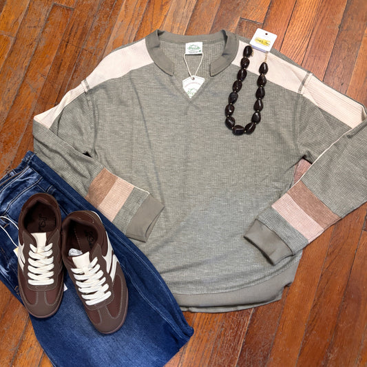 A flat-lay outfit arrangement on a wooden floor featuring a heather gray long-sleeve colorblock shirt with beige and brown panels, folded blue jeans, brown sneakers with white laces, a dark beaded necklace, and a small white tag.