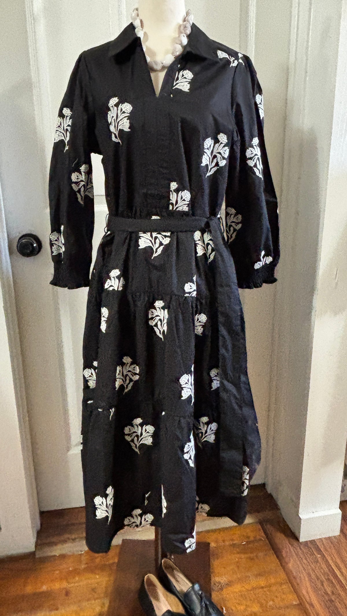 A black long-sleeve midi dress with a white floral print, displayed on a mannequin in an indoor setting with a wooden floor and white door frame.