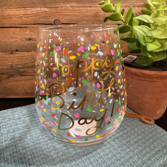 A clear stemless wine glass with gold foil lettering that reads 'Happy Birthday!' and decorated with colorful confetti dots, set on a gray textured cloth with a wooden wall and a potted succulent in the background.