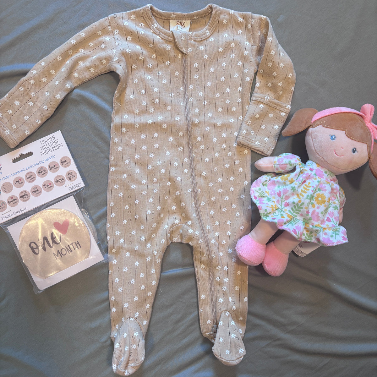 A baby footed sleeper in beige with white daisy patterns, laid flat on a gray fabric background next to a 'One Month' hat in its packaging and a plush doll.