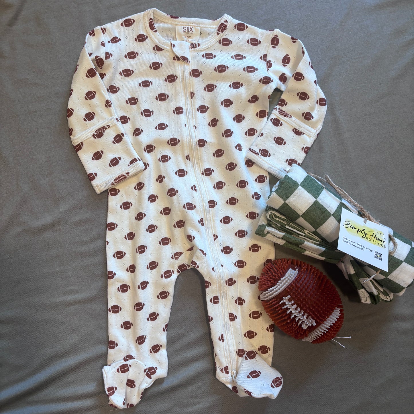 A baby footed one-piece sleeper in cream with a brown football print, laid flat on a gray fabric background next to a green and white checkered blanket and a brown football-shaped toy. The brand tag reads Simply The Thing.