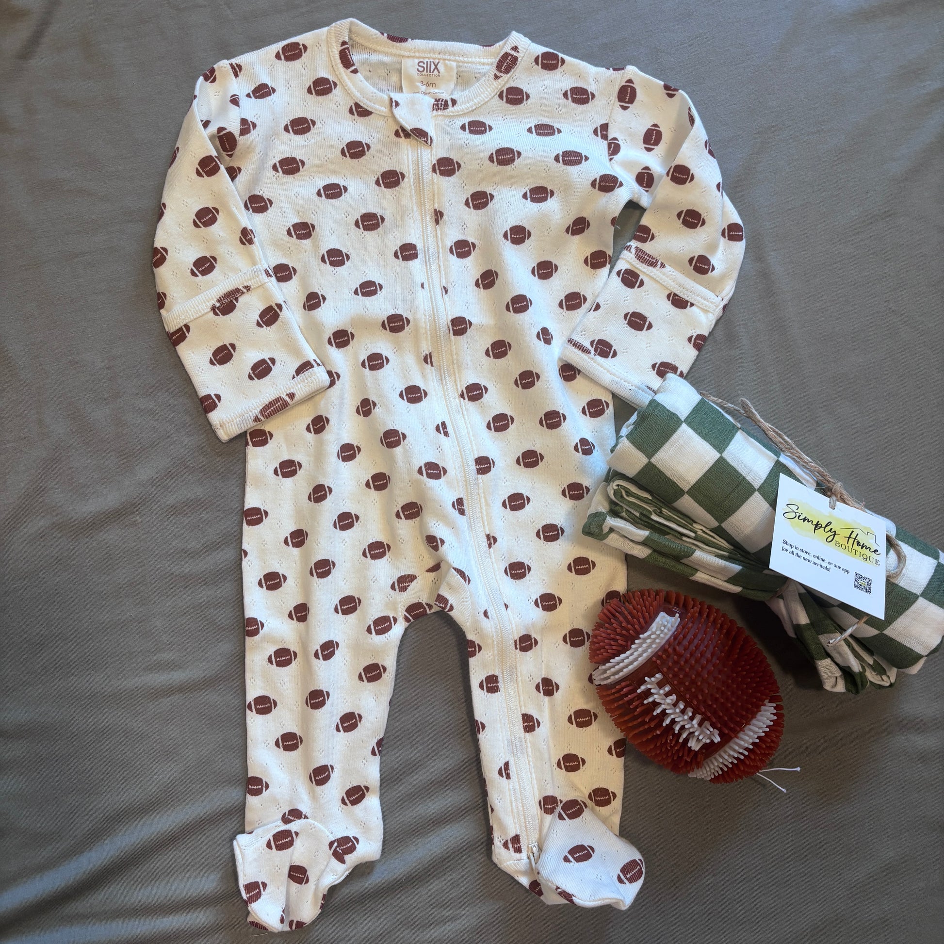 A baby footed one-piece sleeper in cream with a brown football print, laid flat on a gray fabric background next to a green and white checkered blanket and a brown football-shaped toy. The brand tag reads Simply The Thing.
