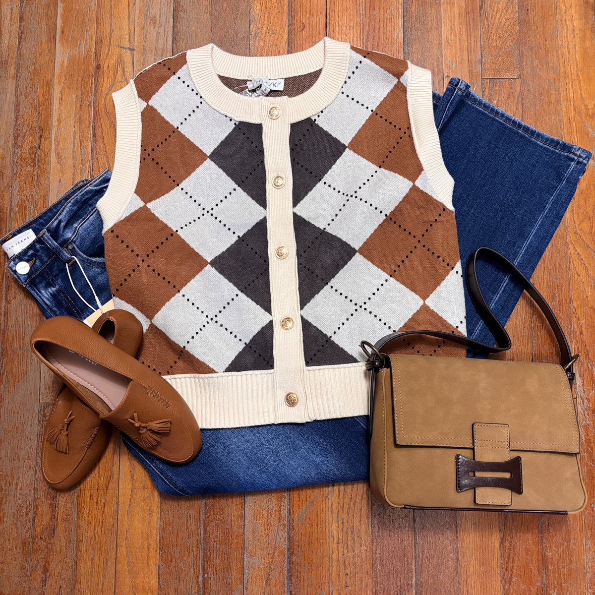 A styled flat-lay outfit on a wood floor featuring a brown, beige, and gray argyle knit cardigan vest, folded blue jeans, brown tassel loafers, and a brown leather handbag.