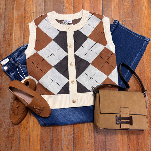 A styled flat-lay outfit on a wood floor featuring a brown, beige, and gray argyle knit cardigan vest, folded blue jeans, brown tassel loafers, and a brown leather handbag.