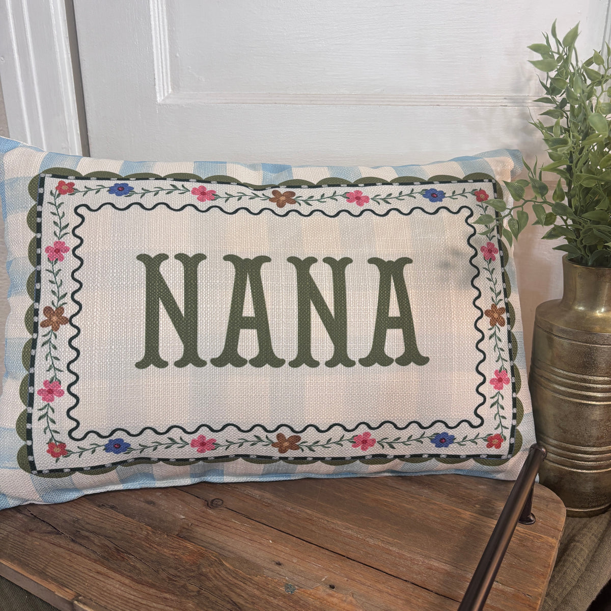 A rectangular decorative pillow with the word “NANA” in large dark green letters, surrounded by a floral embroidered border, resting on a rustic wooden bench in front of a white door with a potted plant nearby.