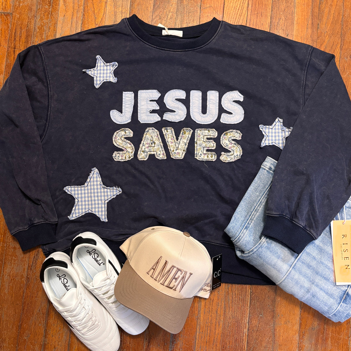 A dark navy crewneck sweatshirt with 'JESUS SAVES' and checkered stars, styled in a flat lay on a wooden floor with white sneakers, a beige 'AMEN' cap, and light blue jeans.
