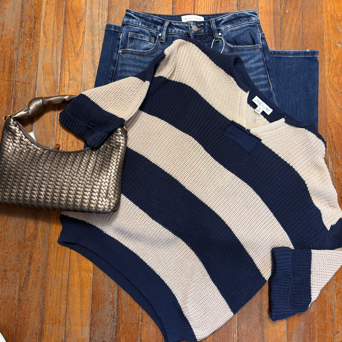 A navy blue and cream striped knit sweater is laid flat on a wooden floor, styled with dark blue jeans and a metallic woven handbag.