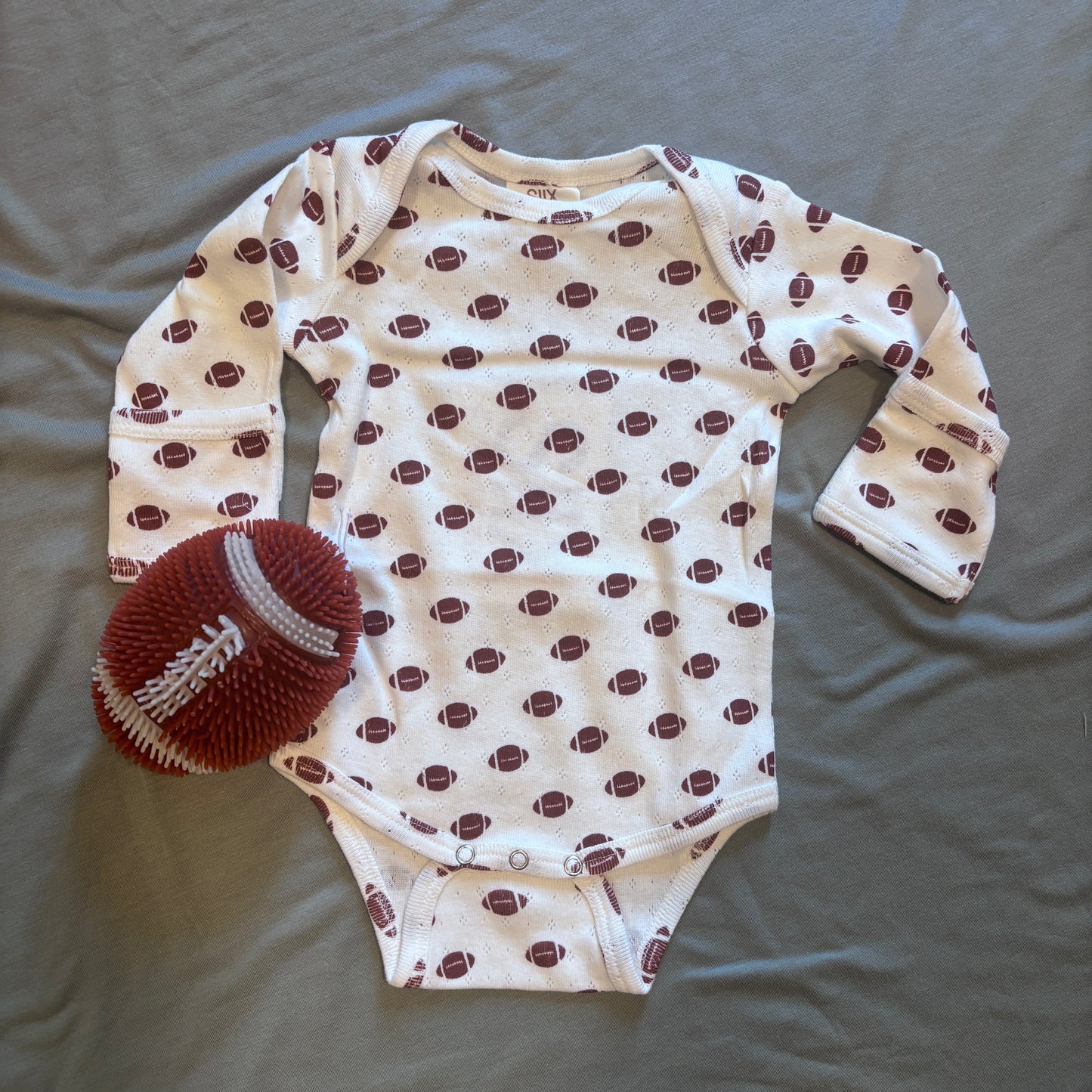 A baby long-sleeve bodysuit in off-white with a repeating pattern of brown footballs, laid flat on a gray fabric background next to a knitted brown football toy.
