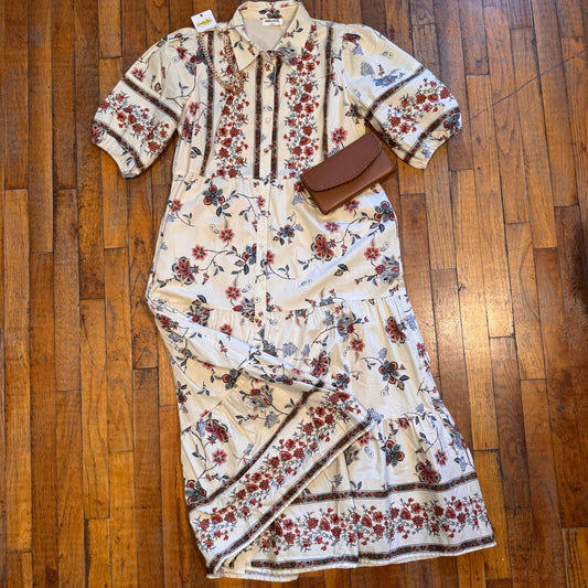 A cream-colored tiered maxi dress with a red and blue floral print, styled with a brown leather clutch on a polished wooden floor.