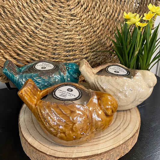 Three ceramic bird-shaped soap bars on a wooden stand with a woven basket and flowers in the background.