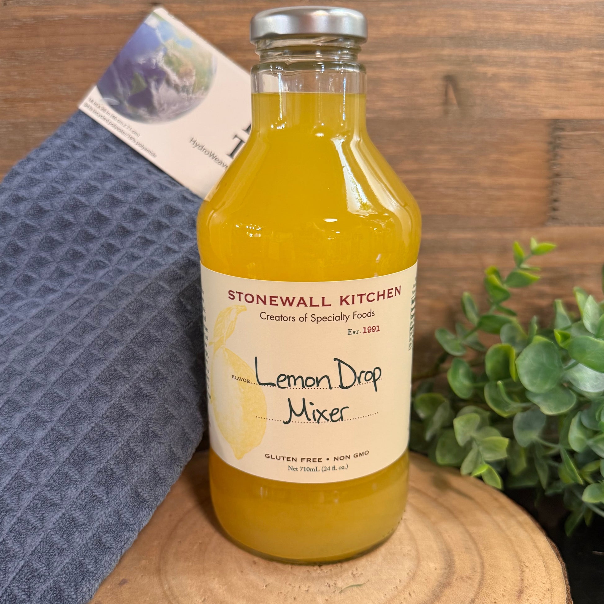 Bottle of Stonewall Kitchen Lemon Drop Mixer on a wooden surface with a towel and plant in the background.