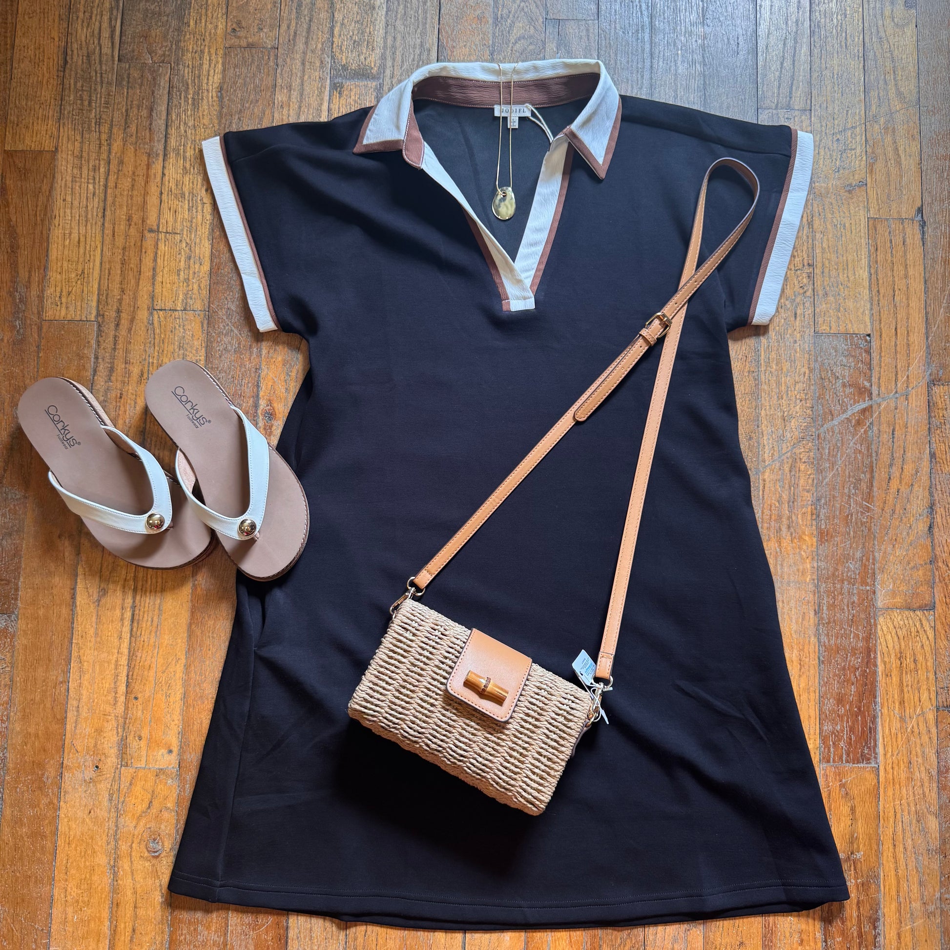 Black dress with white trim, brown sandals, and a woven handbag on a wooden floor.