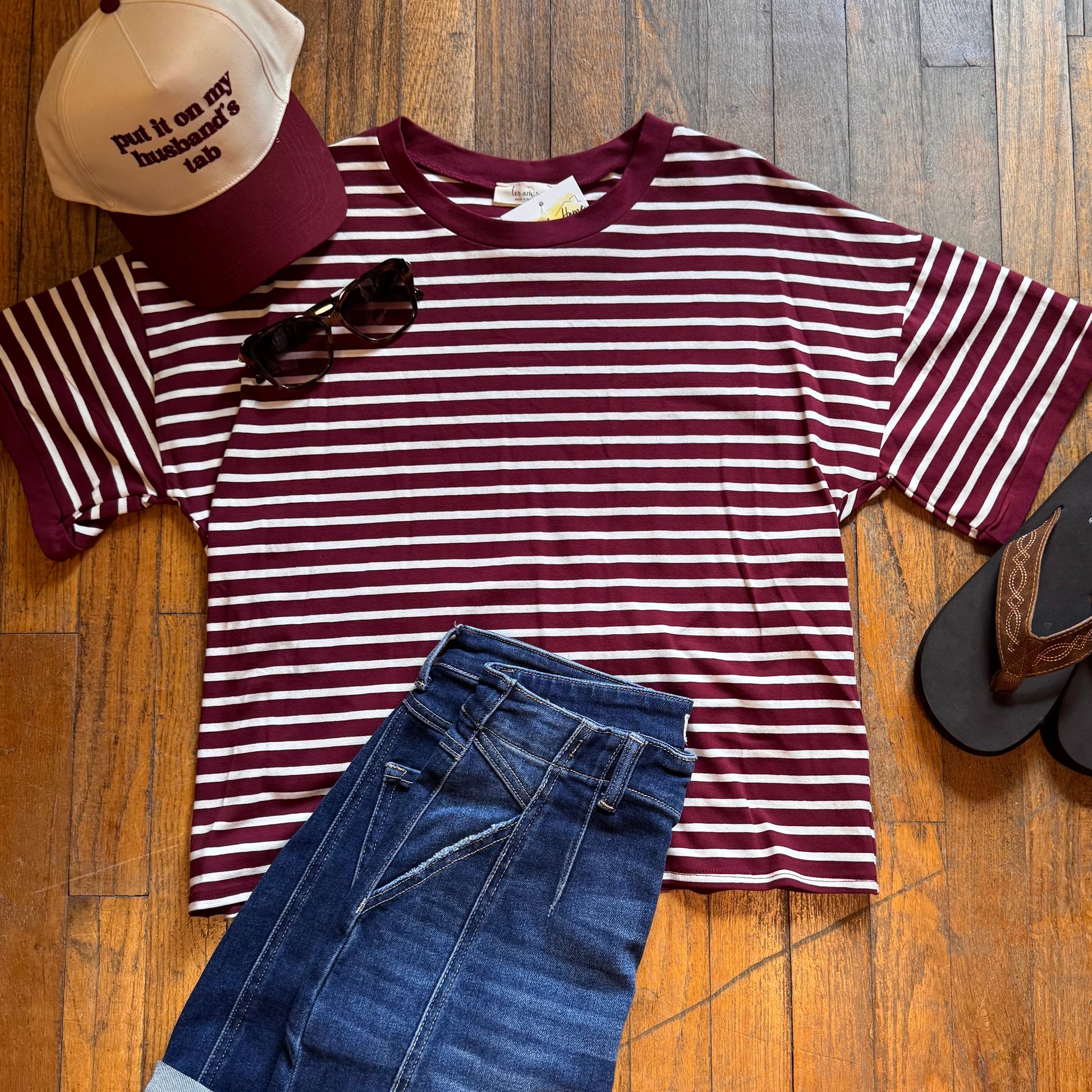 Maroon and white striped shirt with blue jeans, cap, sunglasses, and sandals on a wooden floor.