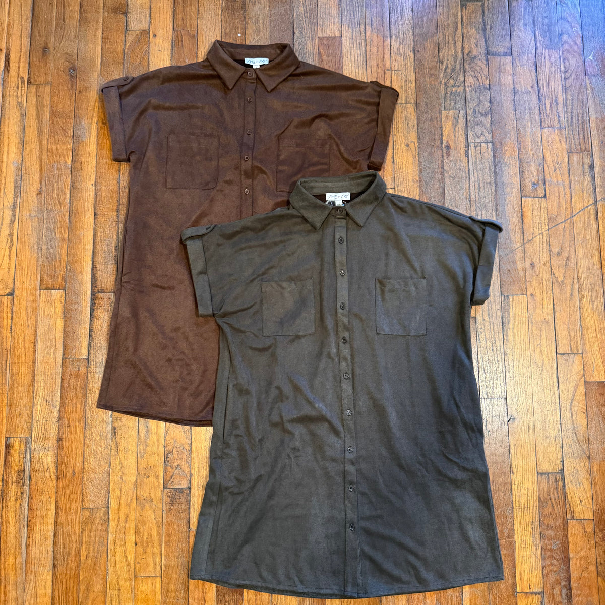 Two short-sleeve button-front collared shirts laid flat on a wooden floor. The top shirt is dark brown, and the bottom shirt is very dark charcoal gray.