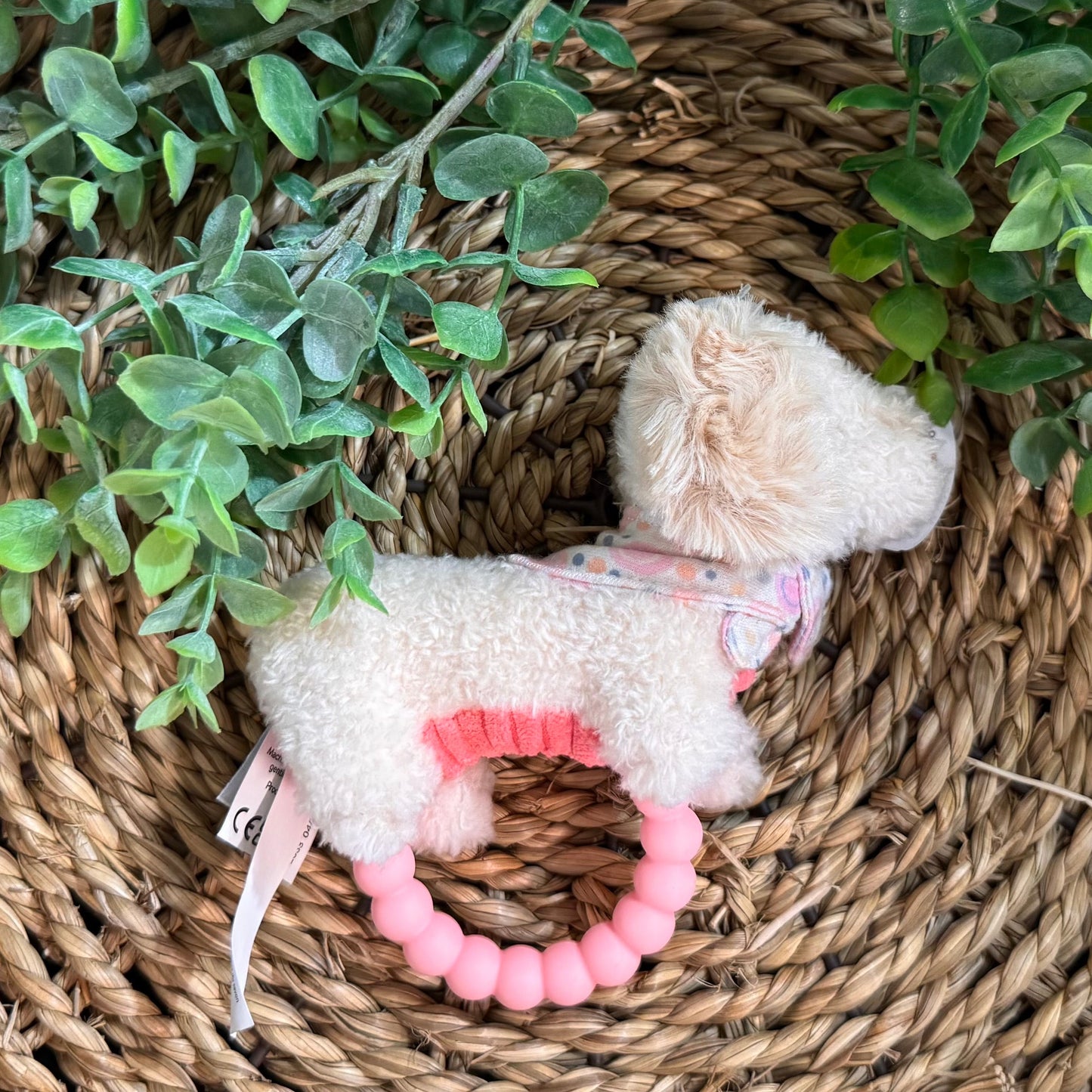 A plush dog-shaped baby teether with a soft cream-colored body, pink ring, and pastel patterned harness, styled on a woven wicker mat with green foliage.