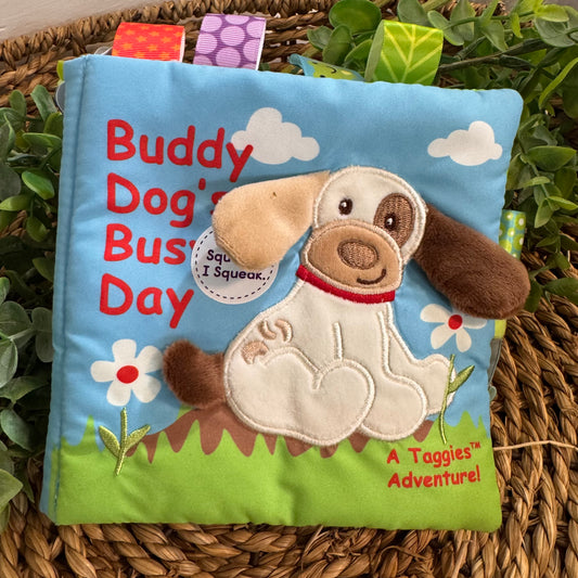 A baby book-style plush toy with a blue cover, featuring an embroidered white dog with brown ears and paws, red text reading 'Buddy Dog's Busy Day,' and a green grassy base. The toy is placed on a woven wicker tray with green leaves around it.