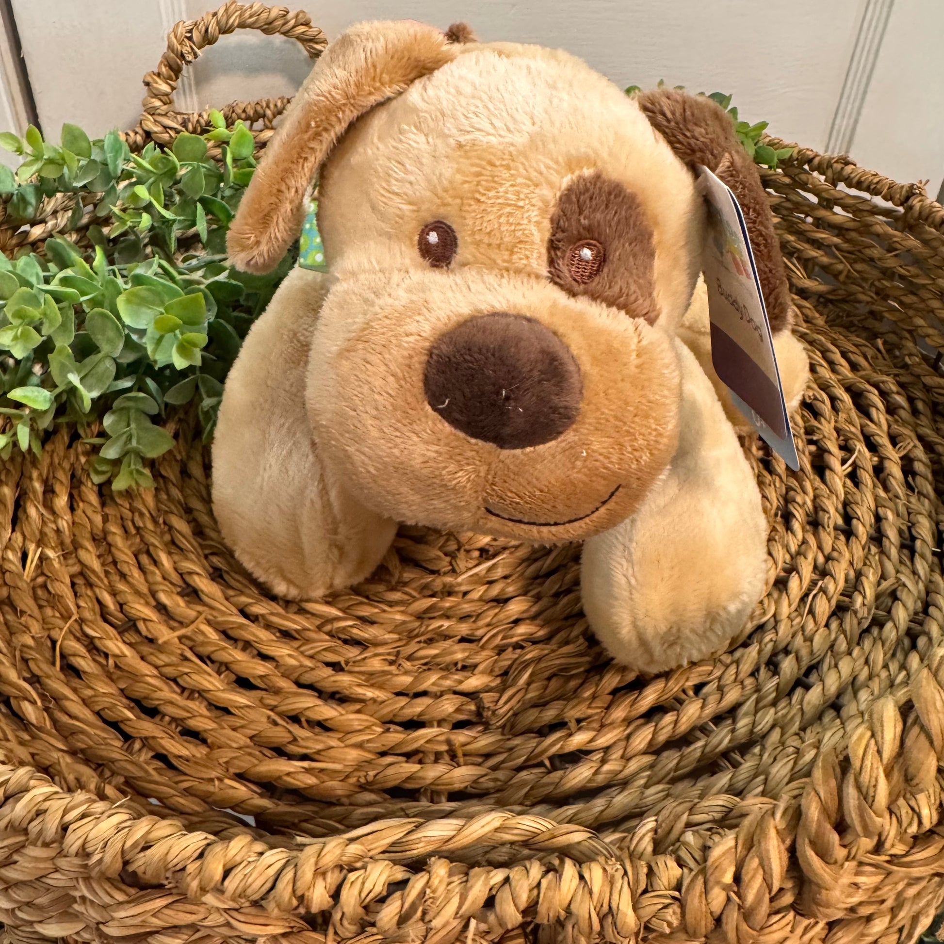 A plush puppy toy in tan and brown, sitting inside a woven straw basket with green leaves in the background.
