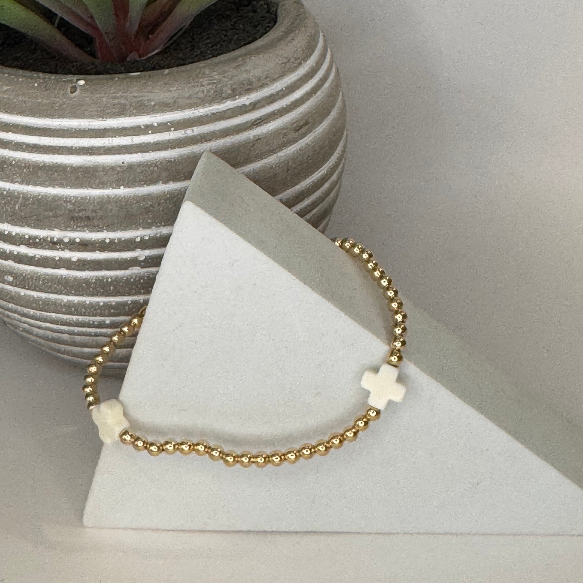 A delicate gold beaded bracelet with two white cross charms, displayed on a white geometric stand next to a potted succulent.