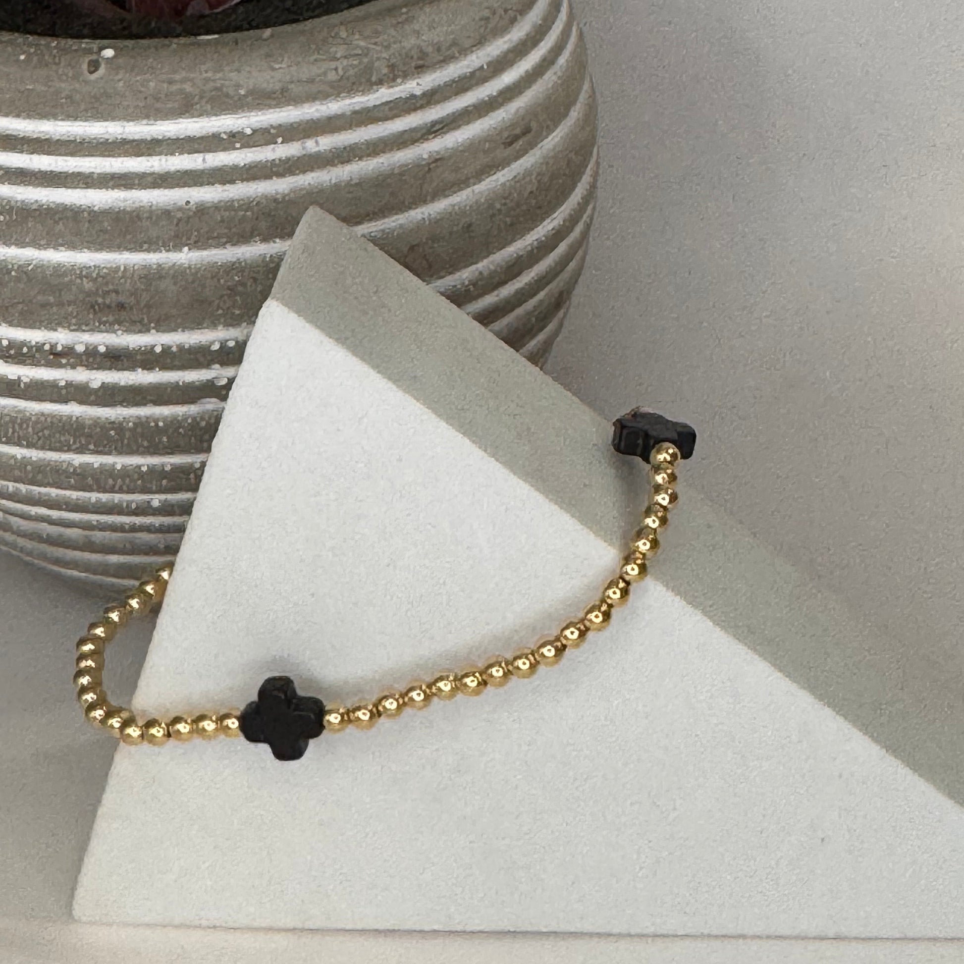 A delicate gold beaded bracelet with black clover-shaped charms, displayed on a white triangular stand next to a potted succulent.