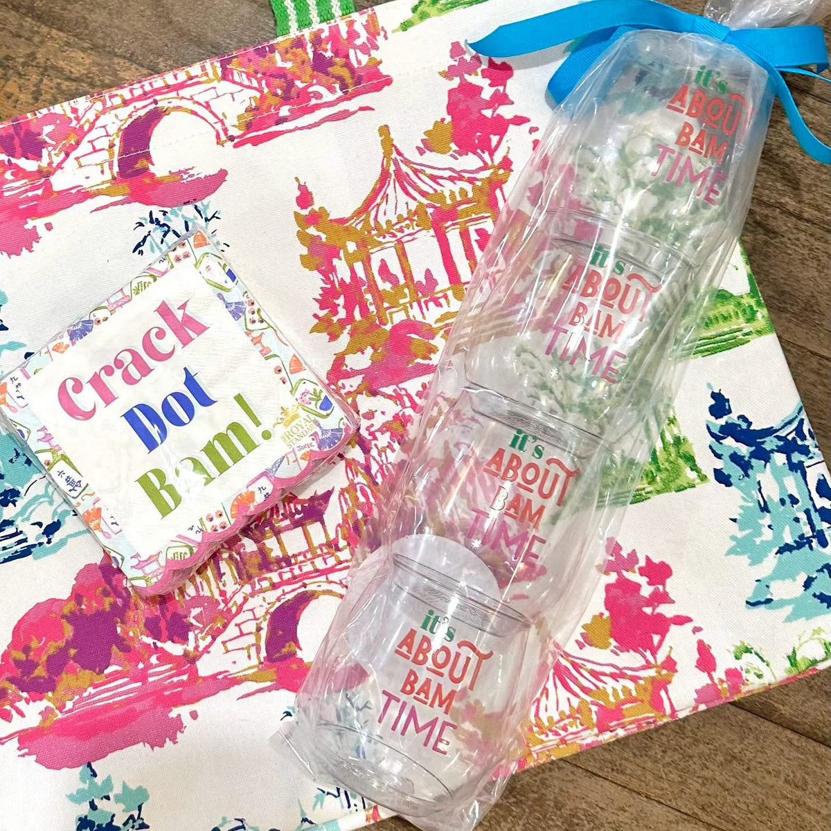 A tote bag with a colorful architectural print and green striped handles, shown on a wooden surface with a small card reading 'Crack Dot Bon!' and a stack of clear plastic cups tied with a blue ribbon.