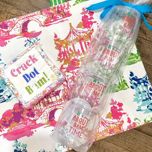 A tote bag with a colorful architectural print and green striped handles, shown on a wooden surface with a small card reading 'Crack Dot Bon!' and a stack of clear plastic cups tied with a blue ribbon.