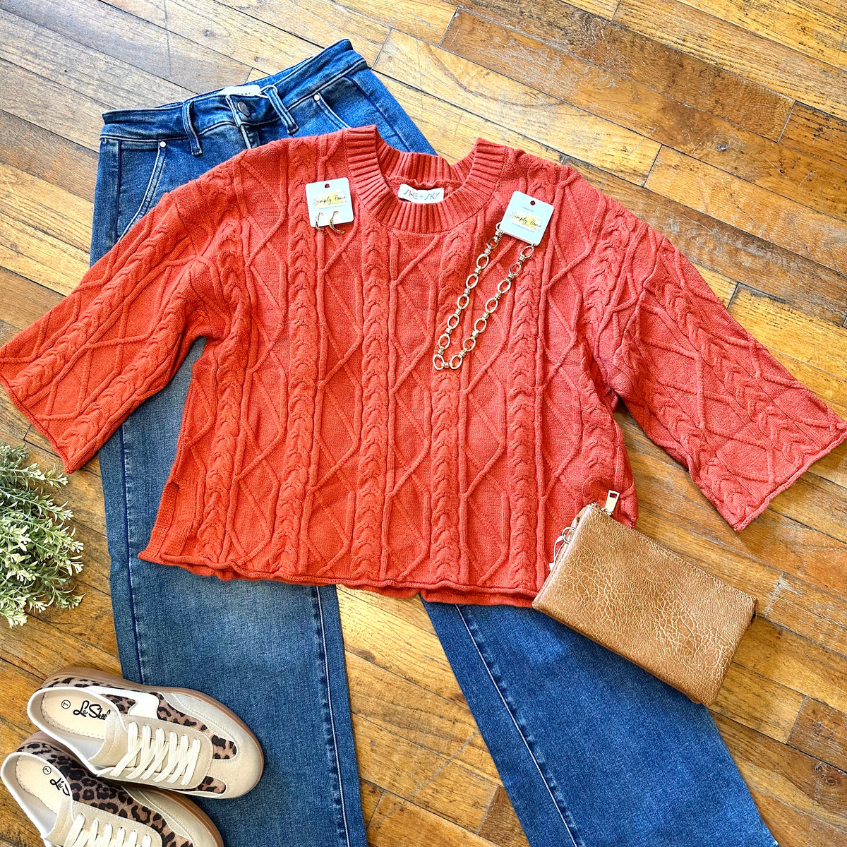 A flat lay of a coral orange cable-knit sweater with three-quarter sleeves, styled over blue jeans on a wooden floor, accessorized with a gold chain necklace, a brown clutch, and beige-and-cream sneakers.