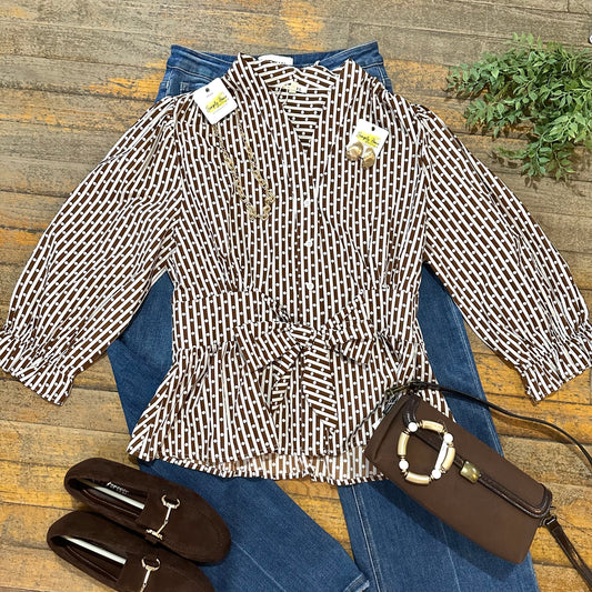 A women's long-sleeve belted blouse with a brown and white geometric pattern, styled in a flat lay on blue jeans with a brown handbag and brown loafers on a rustic wooden background.
