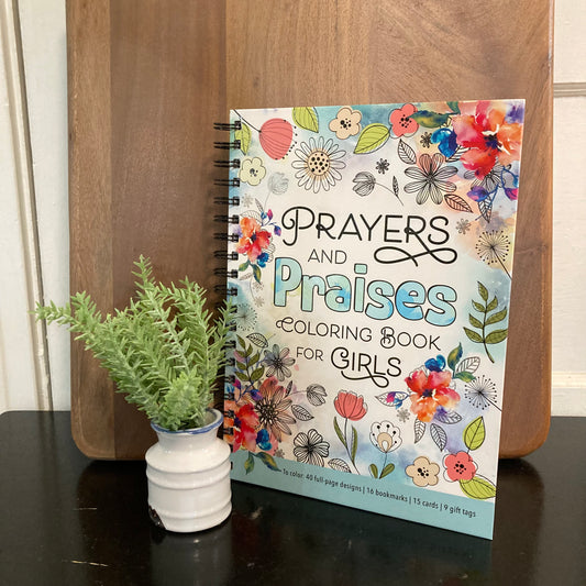 Prayers and Praises Coloring Book for Girls with a colorful floral cover, displayed on a dark table next to a small potted green plant in front of a wooden background.