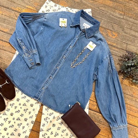 A blue denim long-sleeve button-front shirt laid flat on a wood floor, styled with cream floral pants, a brown leather clutch, and brown shoes. The shirt has pleated details on the shoulders and sleeves, a pointed collar, and a gold-tone chain detail across the chest.