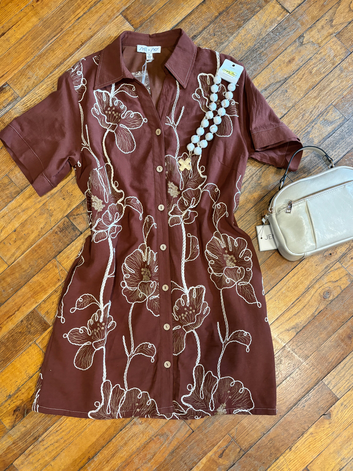 A short-sleeve button-front shirt dress in dark brown with a white floral print, laid flat on a wooden floor next to a cream-colored crossbody bag and a beaded necklace.