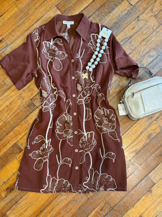 A short-sleeve button-front shirt dress in dark brown with a white floral print, laid flat on a wooden floor next to a cream-colored crossbody bag and a beaded necklace.
