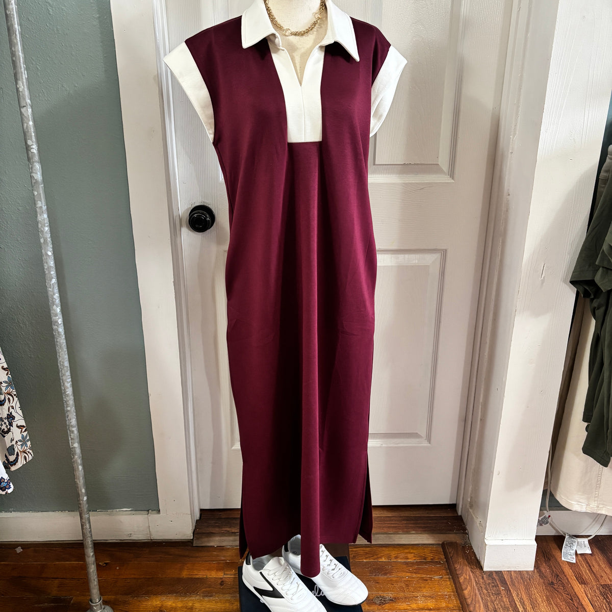 A sleeveless burgundy maxi dress with a white collar and short white sleeves, displayed on a mannequin in front of a white paneled door.