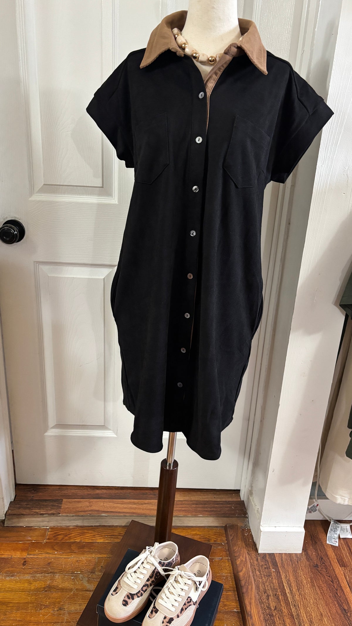 A black short-sleeve button-front shirt dress displayed on a mannequin indoors, with a tan collar and a beaded necklace on the mannequin. A pair of white sneakers sits on the stand below.