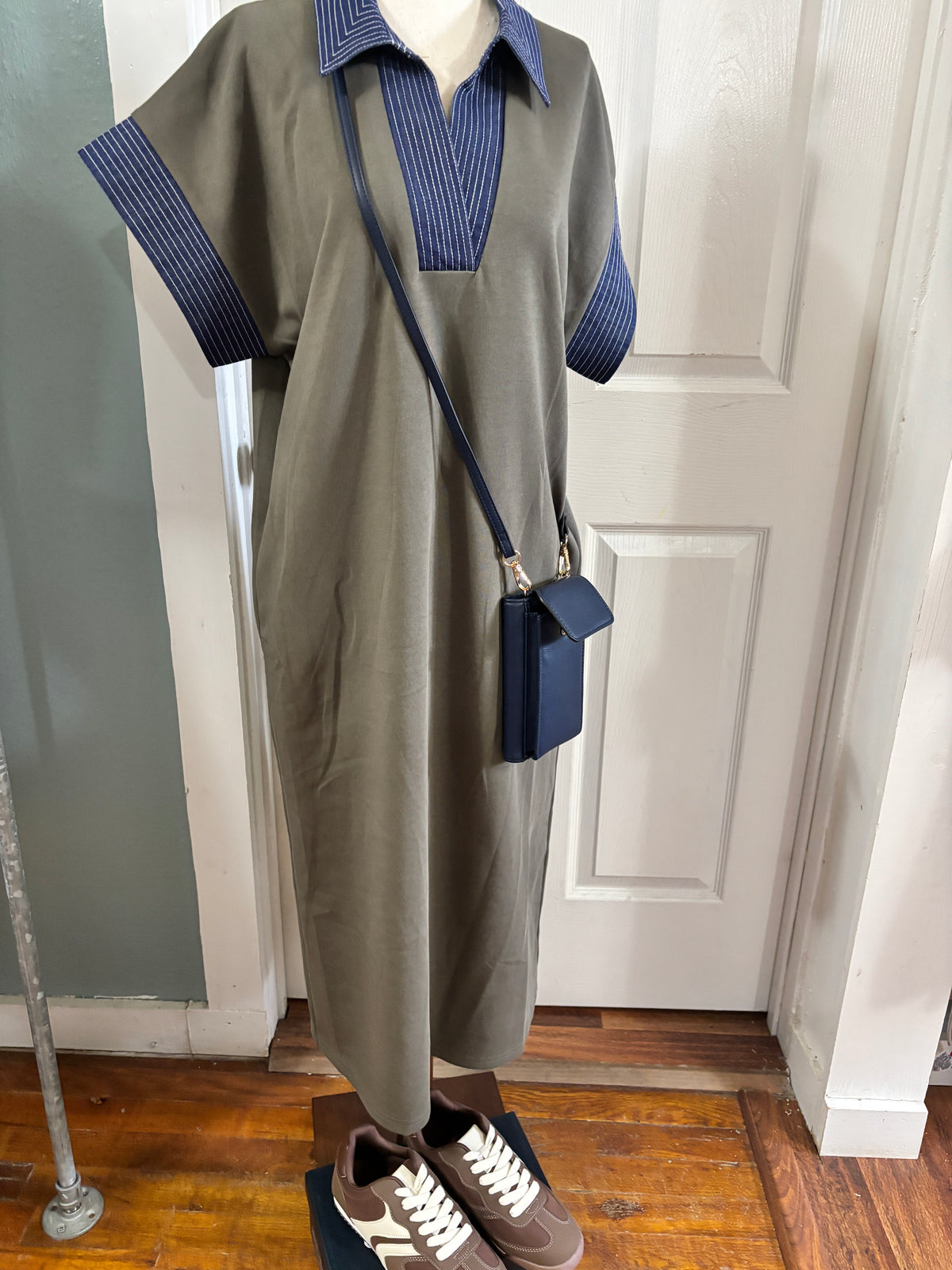 A short-sleeve olive green dress with a blue and white striped V-neck collar and sleeve cuffs, styled on a mannequin with a navy blue crossbody bag and brown sneakers.