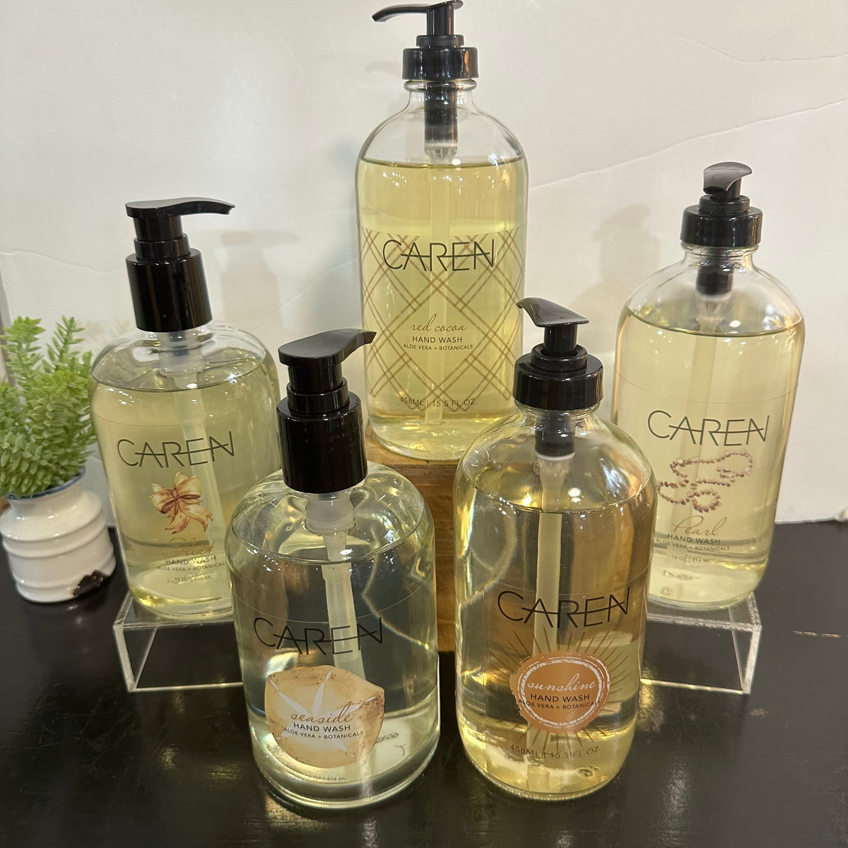 Five Caren hand wash bottles in different sizes arranged on clear acrylic stands, with a small potted plant on the left, set against a white wall and dark tabletop.