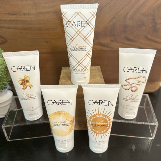 Four Caren hand treatment tubes arranged on clear acrylic stands against a wooden background. The white tubes feature different decorative labels: a gold bow, a gold plaid pattern, a gold sunburst, and a gold leaf design.