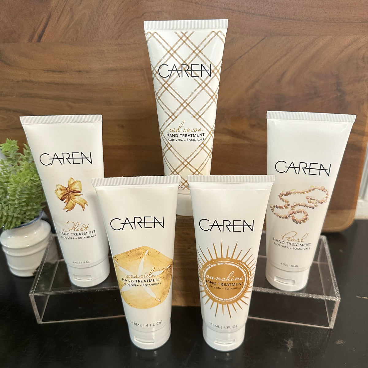 Five Caren hand treatment tubes arranged on a clear acrylic stand on a dark tabletop, with a wooden wall and small potted plants in the background.