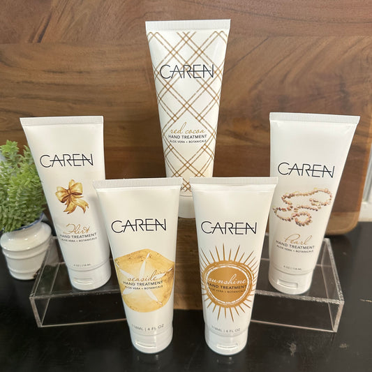Five Caren hand treatment tubes arranged on a clear acrylic stand on a dark tabletop, with a wooden wall and small potted plants in the background.